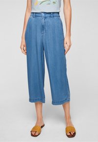 Woman wearing relaxed-fit cropped blue jeans with a buttoned elastic waistband and tan woven slide sandals, standing on a gray background.