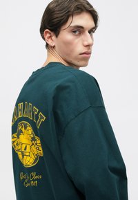 Dark green sweatshirt with a crew neck, featuring a yellow globe graphic and "Carhartt" text on the back. Soft cotton material.