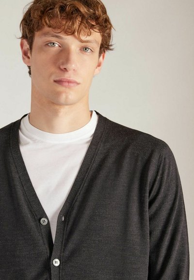 Donkergrijze cardigan met V-hals, gemaakt van zachte stof, voorzien van een knoopsluiting en een gladde textuur, die over een wit T-shirt is gedragen.