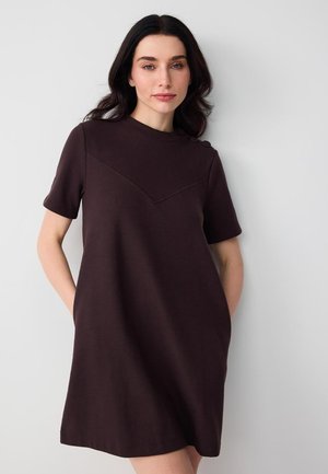 Femme aux cheveux foncés portant une robe noire à manches courtes avec des boutons sur l'épaule, posant devant un fond clair uni.