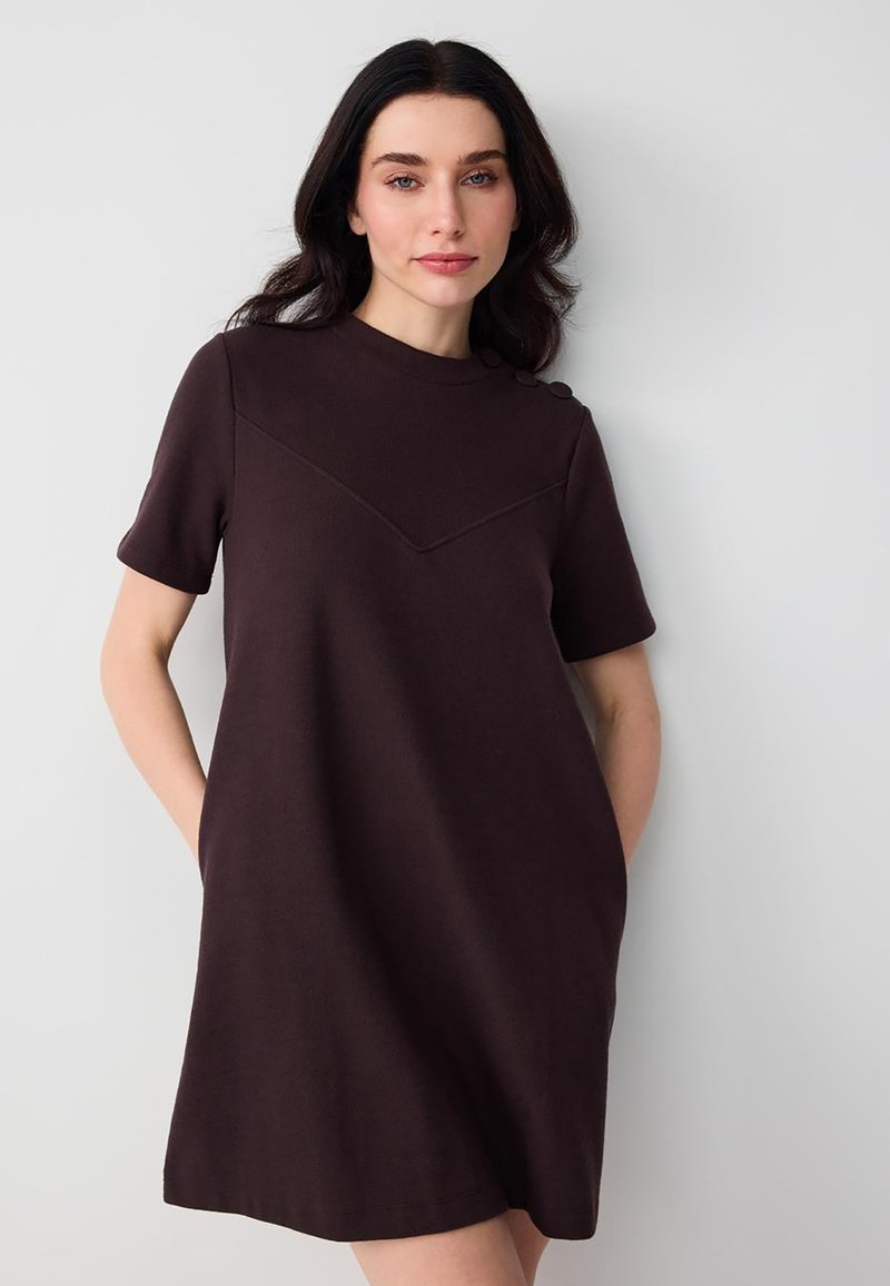 Frau mit dunklem Haar trägt ein schwarzes kurzes Kleid mit Knöpfen an der Schulter und posiert vor einem schlichten hellen Hintergrund.