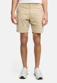 Beige bomullshorts har flat front, sidelommer og en skreddersydd passform, sammen med hvite og grå sneakers.