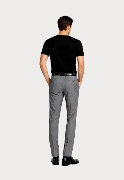 Άνδρας που φοράει μαύρο κοντομάνικο πουκάμισο, γκρι ραμμένο παντελόνι με μαύρη ζώνη και μαύρα επίσημα παπούτσια, στέκεται με τα χέρια στις τσέπες, γυρισμένος προς τα πίσω.