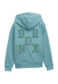 Huvtröja i ljusblått med strukturerade, glittriga bokstäver som stavar "BRONX" på baksidan. Har en avslappnad passform och ribbade ärmslut.
