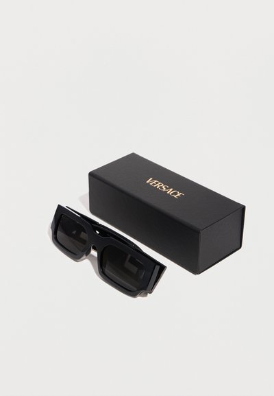 Μαύρα ορθογώνια γυαλιά ηλίου δίπλα σε μαύρη θήκη Versace με γράμματα σε χρυσό χρώμα πάνω σε λευκή επιφάνεια.
