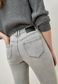 Persona vistiendo jeans de mezclilla gris claro y un suéter de canalé gris, con una mano metida en un bolsillo trasero.