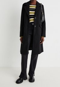 Abrigo de lana negro con grandes solapas, suéter de rayas verdes y blancas con canalé, jeans negros y botas negras robustas. Acompañado de un bolso pequeño.