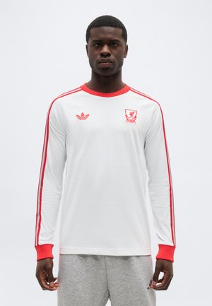 Mees, kes kannab valget pika varrukaga särki, millel on punane ääris, punased triibud varrukatel ja Liverpooli jalgpalliklubi embleem, seisab tavalise tausta ees.