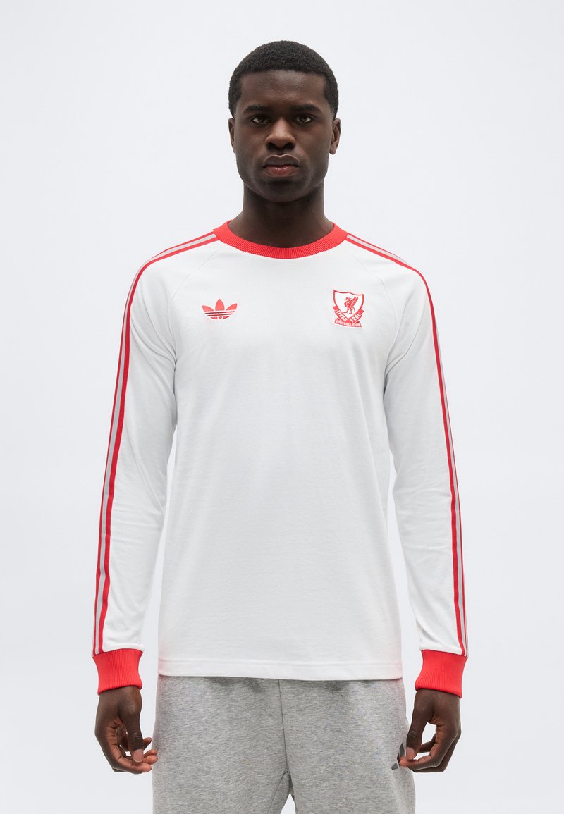 Vyras dėvi baltą ilgomis rankovėmis marškinį su raudona apdaila, raudonomis juostelėmis ant rankovių ir Liverpulio futbolo klubo herbu, stovintis priešais paprastą foną.