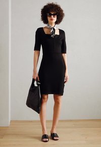 Robe noire ajustée avec des manches courtes, décolleté carré, en tissu lisse. Associée à des sandales glissantes noires et un sac noir. Écharpe blanche à motifs comme accent.