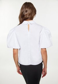 Weiße Bluse mit puffähnlichen Ärmeln, die an der Rückseite mit einem Schlüssellochverschluss versehen ist und einen strukturierten Kragen hat, aus glattem Baumwollstoff gefertigt.