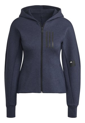 Person iført en mørkeblå hættetrøje med lynlås og matchende bukser med tre lodrette sorte striber på brystet og låret, hænderne i lommerne.