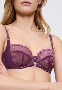 Fialová krajková podprsenka s průsvitnými košíčky se květinovými vzory, dekorativním kroužkem uprostřed a nastavitelnými ramínky.