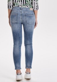 Jeans ajustados en denim azul claro, con una cinturilla alta, dos bolsillos traseros y un dobladillo ligeramente desgastado. Combinados con zapatillas blancas.