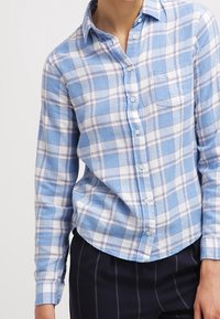 Modrá a bílá kostkovaná flanelová košile s předním zapínáním na knoflíky, límečkem, jednou náprsní kapsou a mírně zaobleným spodním lemem. Texturovaná tkanina.
