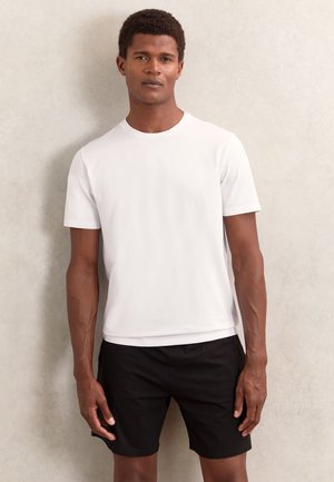 Ung mand står op ad en lys væg iført en enkel hvid t-shirt med korte ærmer og sorte shorts, kigger lige frem.