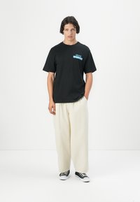 Sort kortærmet t-shirt med et blåt grafisk logo på brystet, parret med cremefarvede vidde bukser og sort-hvide sneakers.