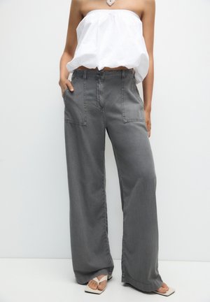 Frau trägt lockere graue Hose mit Vordertaschen, ein weißes trägerloses Top, beige Sandalen mit quadratischer Spitze und eine Halskette in Herzform.