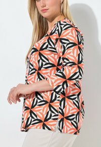 Mujer con cabello rubio largo, vestida con una blusa de patrón geométrico en naranja, negro y blanco, con mangas hasta tres cuartos, posando de perfil.