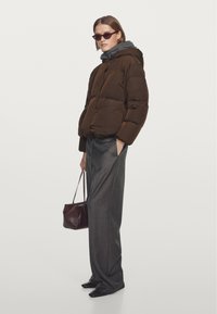 Brun dunjakke med grå hætte, båret sammen med grå bukser med vide ben. Modellen bærer en lille mørk taske og har mørke solbriller på.