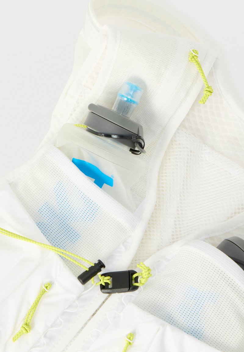 Gilet idratante in rete bianca con una borraccia morbida in una tasca frontale, cordini gialli e chiusure con fibbie in plastica nera.