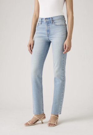 Femme portant un jean bleu clair à jambes droites, un top blanc sans manches et des sandales beige à talons bas avec des brides, debout devant un fond uni.