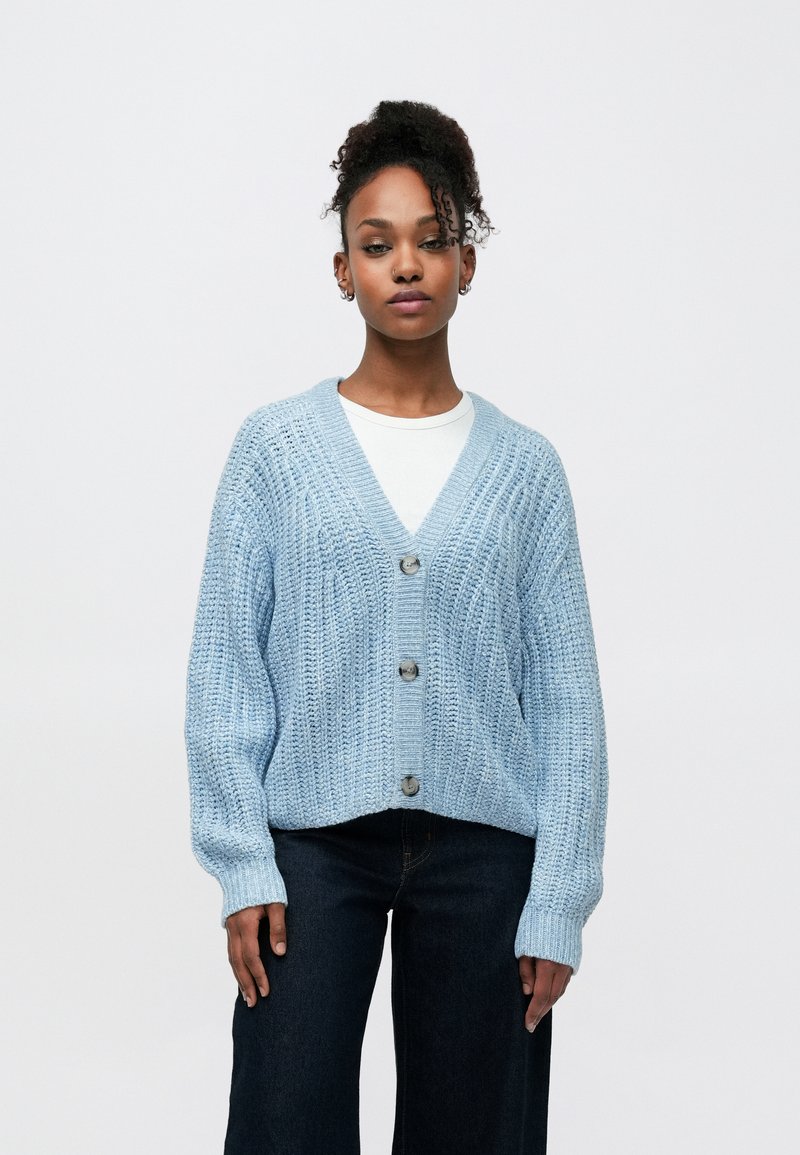 Hellblauer gestrickter Cardigan mit V-Ausschnitt, fünf Knöpfen und strukturiertem Muster. Kombiniert mit einem weißen Top und dunklen, weit geschnittenen Jeans.