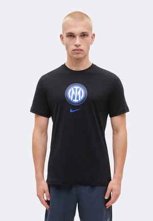 Ung mand med kort blondt hår iført sort t-shirt med blåt og hvidt sportslogo og blå Nike-swoosh, står mod en ensfarvet baggrund.