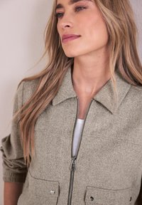 Frau mit langen blonden Haaren, die eine hellgraue Jacke mit Reißverschluss über einem weißen Oberteil trägt, leicht seitlich stehend mit neutralem Gesichtsausdruck.