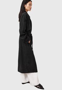 Svart trenchcoat med mjuk mocka-textur, stor krage, vida ärmar och ett bälte. Bärs med vita vida byxor och svarta skor.