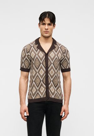 Noor mees, kes kannab lühikeste varrukatega nööpidega särki pruunide ja beežide teemantmustritega, koos mustade pükstega, seisab ühevärvilise tausta ees.