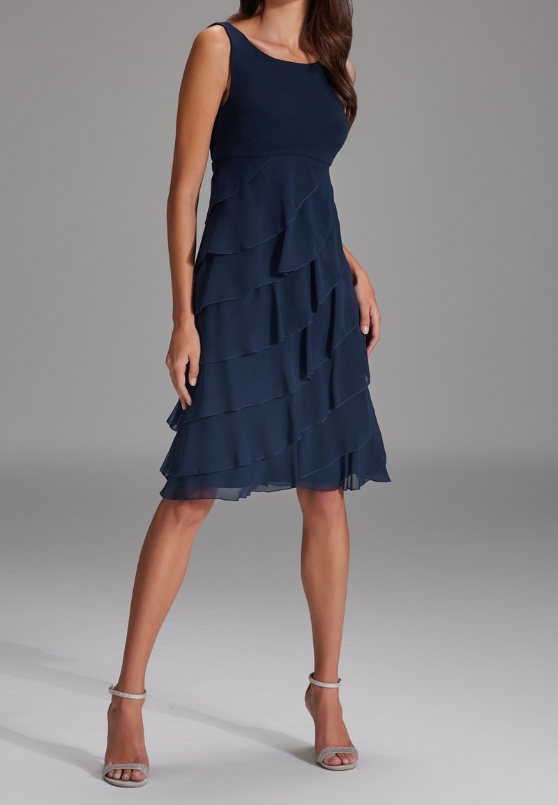 Femme portant une robe bleu marine sans manches, longueur genou, avec des volants superposés et des talons argentés à bride autour de la cheville, debout sur un fond gris.