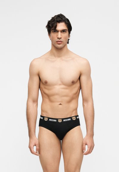Άνδρας χωρίς πουκάμισο με κοντά σγουρά μαλλιά, που φοράει μαύρα εσώρουχα με το λογότυπο Moschino και εικόνες αρκουδάκια στη μέση, μπροστά σε απλό φόντο.