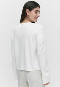 Witte geribbelde long-sleeve top met een ronde halslijn en een kleine metalen accent aan de achterkant. Ontspannen pasvorm, zachte textuur en een iets cropped lengte.