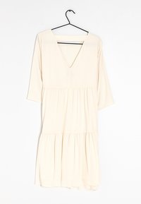 Vestido color crema con escote en V, mangas de tres cuartos y cintura con fruncido, que presenta una falda con volantes y una tela suave y ligera.
