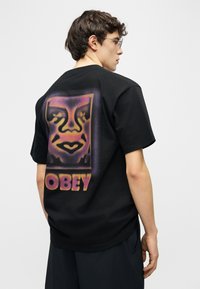 Νεαρός άνδρας με γυαλιά που φοράει μαύρο μπλουζάκι με μεγάλο πορτοκαλί και μοβ στυλιζαρισμένο πρόσωπο και το κείμενο "OBEY" στην πλάτη.