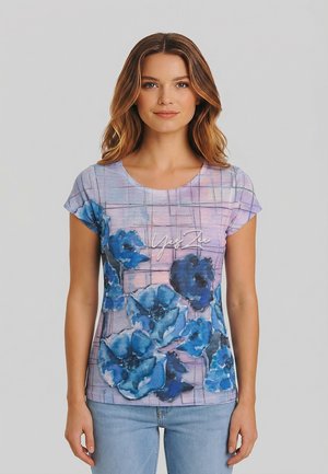 Donna con capelli castani ondulati indossa una maglietta viola a maniche corte con stampa floreale blu e jeans azzurri, in piedi davanti a uno sfondo semplice.