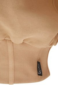 Maglione beige a coste con una vestibilità aderente e orlo arricciato. Presenta un piccolo etichetta nera con dettaglio del logo bianco sul bordo inferiore.