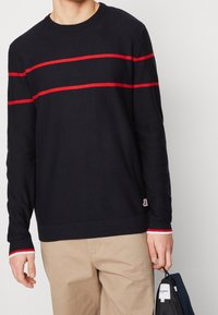 Pull en maille bleu marine avec des rayures rouges horizontales sur la poitrine et les manches. Poignets et ourlet côtelés, avec un patch logo à la taille.