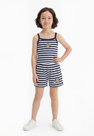 Copil purtând maiou în dungi albastre închise și albe și pantaloni scurți cu petice cu ursuleți de pluș, stând cu mâna pe șold, adidași albi.