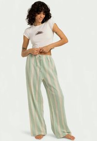 Balta trumpa marškinėliai su maža grafika, derinami su šviesiai žaliais vertikaliai dryžuotais plačiomis kelnėmis, turinčiomis raištelių liemenį.