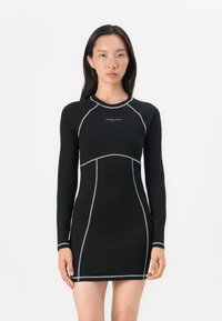 Robe noire à manches longues avec des coutures argentées et une silhouette ajustée, présentant un col rond et un logo de la marque sur la poitrine.