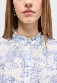 Νεαρή γυναίκα φοράει λευκό πουκάμισο με λεπτομερείς μπλε αγροτικές σκηνές, γιακά με βολάν και μικρά χρυσά και λευκά κουμπιά.