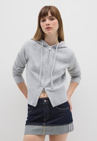 Felpa grigia con zip in morbido maglione a costine, con orlo e polsini a coste. Abbinata a una gonna di jeans scuro con risvolto di jeans chiaro. Design minimalista.
