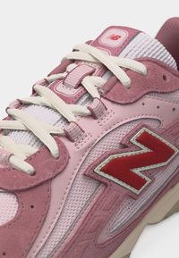 Ροζ και λευκό αθλητικό παπούτσι New Balance με λευκά κορδόνια, κόκκινο λογότυπο "N", υφασμάτινο πλέγμα και σουέντ επικαλύψεις σε κοντινή όψη.