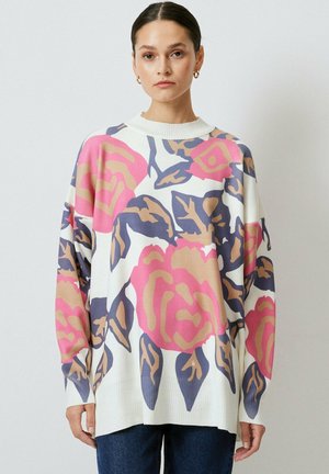 Femme portant un pull blanc ample avec de grands motifs floraux roses et beiges ainsi que des feuilles bleu foncé, associé à un pantalon bleu foncé.