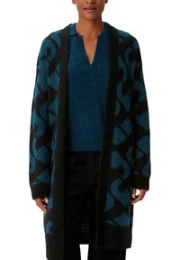 Langer schwarzer Cardigan mit einem geometrischen teal Muster, über einem einfarbigen teal Pullover getragen. Stricktextur mit einem V-Ausschnitt-Design.