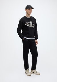 Μαύρο φούτερ με λευκό γραφικό που απεικονίζει βουνά και αναβατήρες σκι, σε συνδυασμό με μαύρη φόρμα και μπεζ αθλητικά παπούτσια, φορεμένα με μαύρο καπέλο.