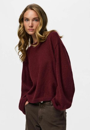 Jeune femme aux cheveux blonds ondulés portant un pull ample bordeaux à manches longues et un pantalon marron foncé, les mains dans les poches.