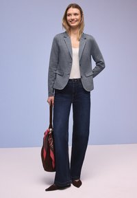 Femme souriante portant un blazer gris, un haut blanc, un jean large sombre, des chaussures pointues, tenant un sac marron avec un foulard rose à motif sur un fond clair.